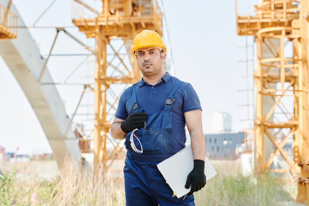 Man in safety gear holding blueprint on construction site with cranes.